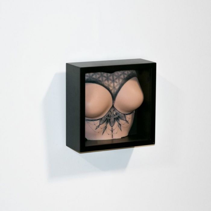 Light-toned tattooable flesh in the shape of female breasts with upper-torso, tattooed with a geometric pattern. Framed in a black box on a white background.