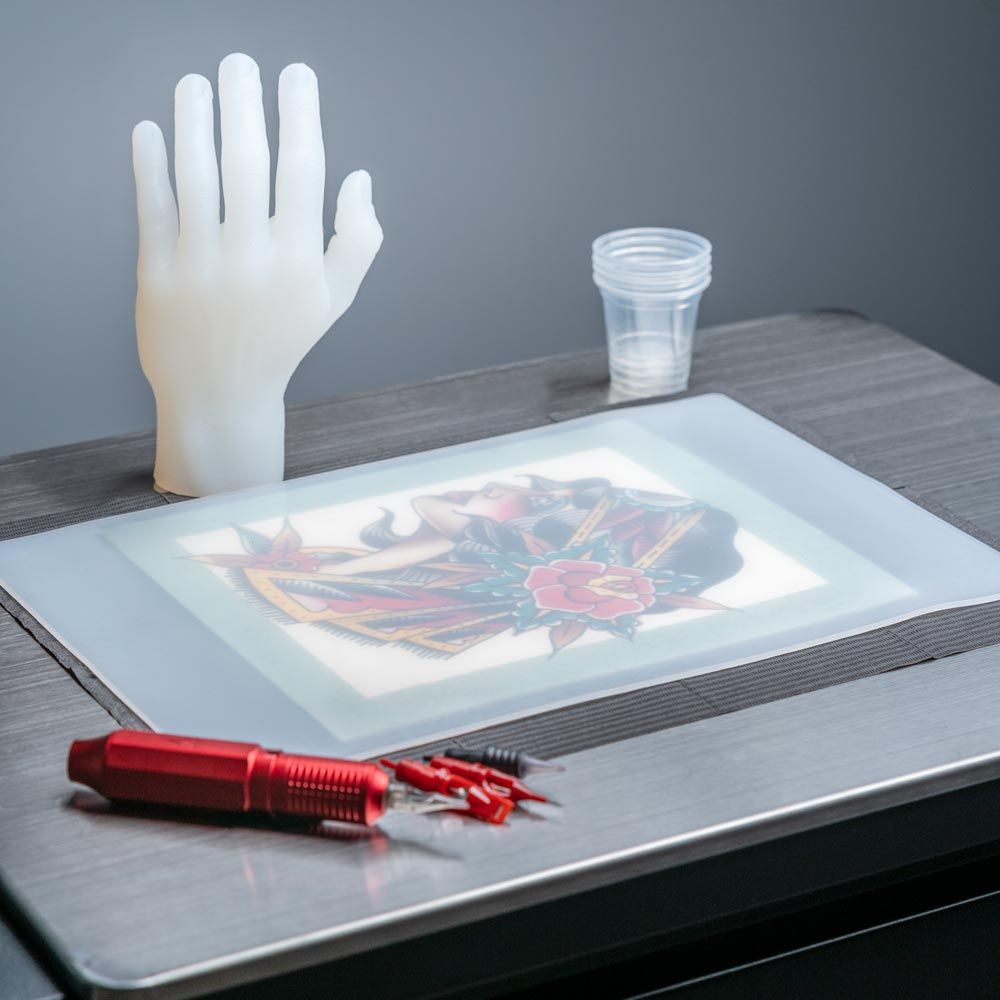 Clear hand made of tattooable flesh laying next to a traditional American woman face tattooed on a rectangle of tattooable flesh on a small table.